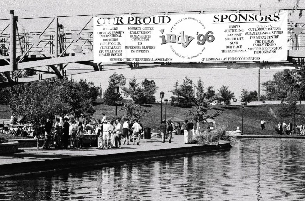 A crowd of people are gathered on the side of a canal. A large sign hangs from a bridge advertising the 1996 Pride festival.