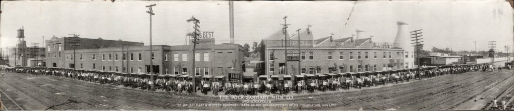 A long row of delivery trucks are parked outside of a factory building. The drivers stand next to their trucks. 