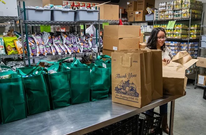 A woman is opening a paper grocery bag. The table next to her is covered in bags full of food. The shelves in the background are filled with grocery items. 