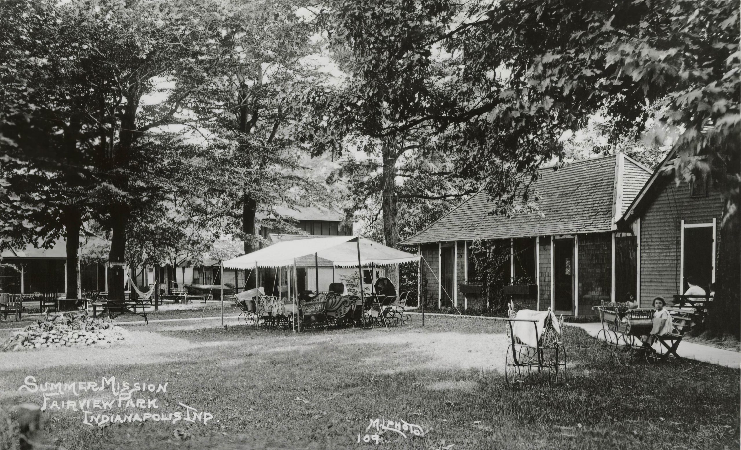 One-story cabins line two sides of a shaded yard. A large white canopy is erected on the grass.