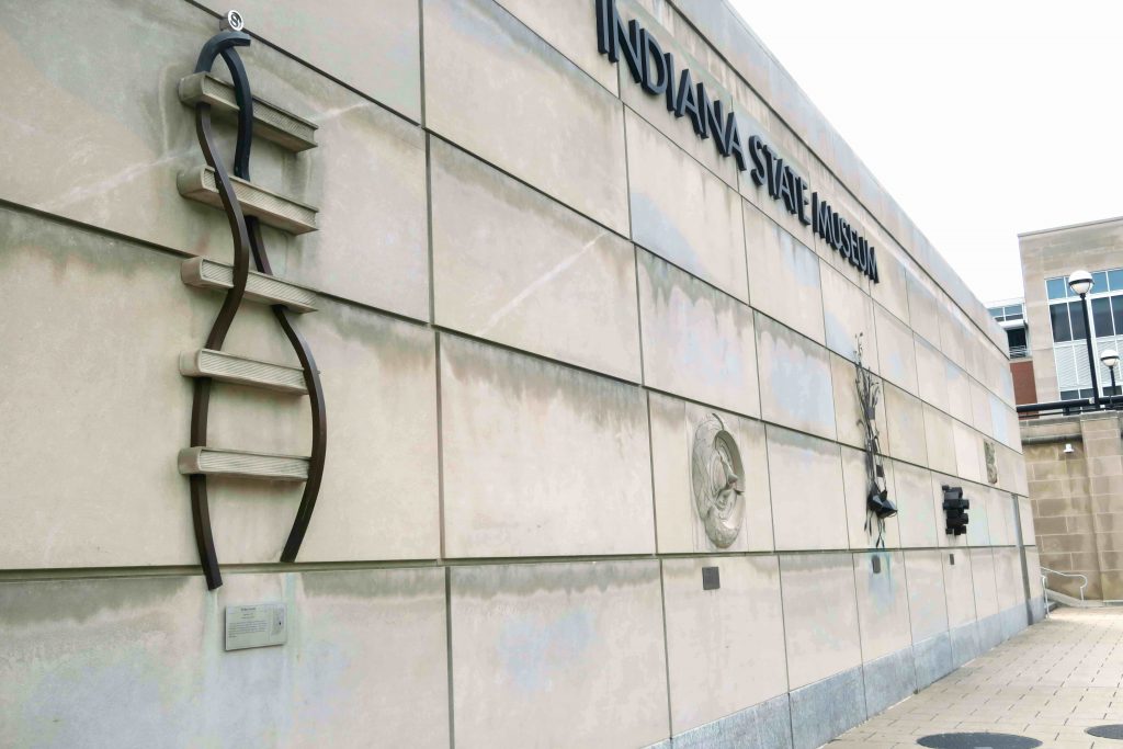 Five piece of art hang on a wall outside. Letters at the top of the wall read Indiana State Museum. 