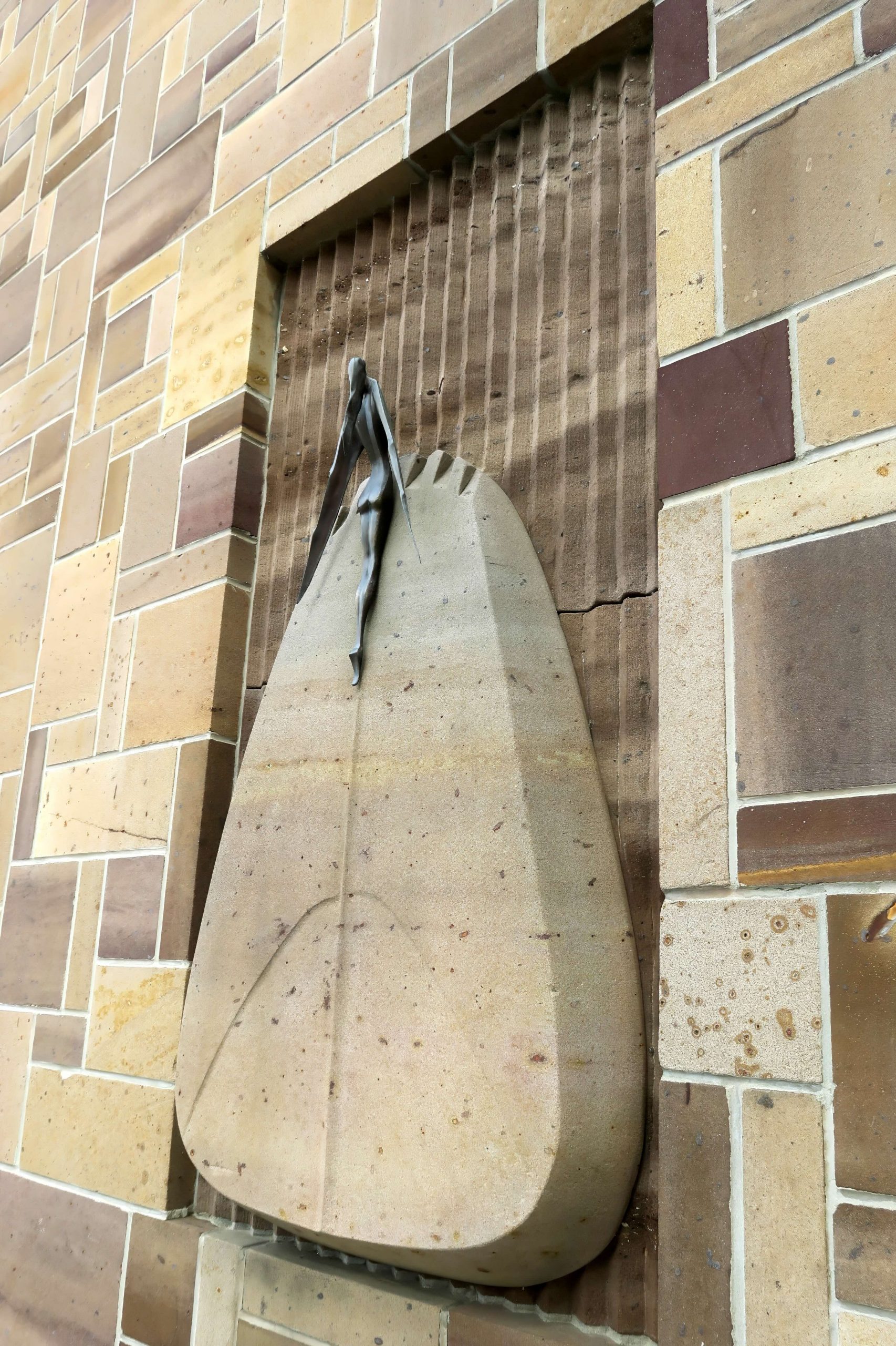 A sculpture on the side of a wall. The piece has a small bronze winged figure affixed to the top of a triangular piece of stone. Behind it is a wave textured section of wall.