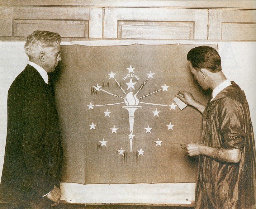 Two men stand in front of a flag. One man holds a sheet of gold leaf and a utensil. 