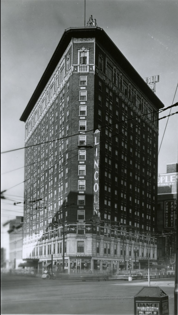 Exterior view of a triangular, 14 story brick building. A large marquee on the side of the building reads Lincoln. The ground level has several store fronts. 