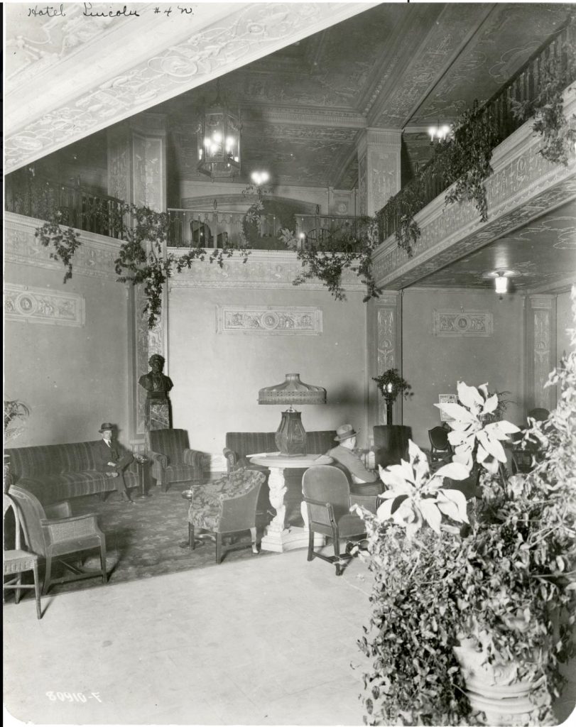 A two-story lobby with a carpeted seating area filled with couches and chairs. Vines hang from the railing on the second floor. Two men sit in chairs. 