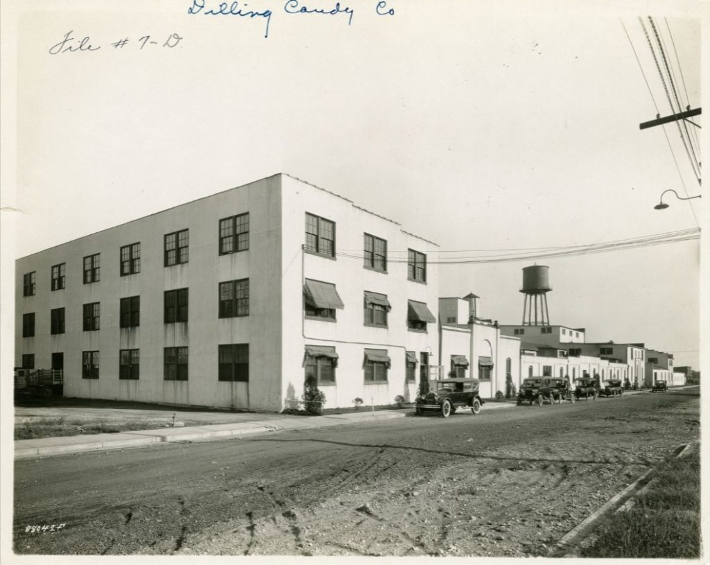 A long factory building with cars parked in front on a dirt road. 