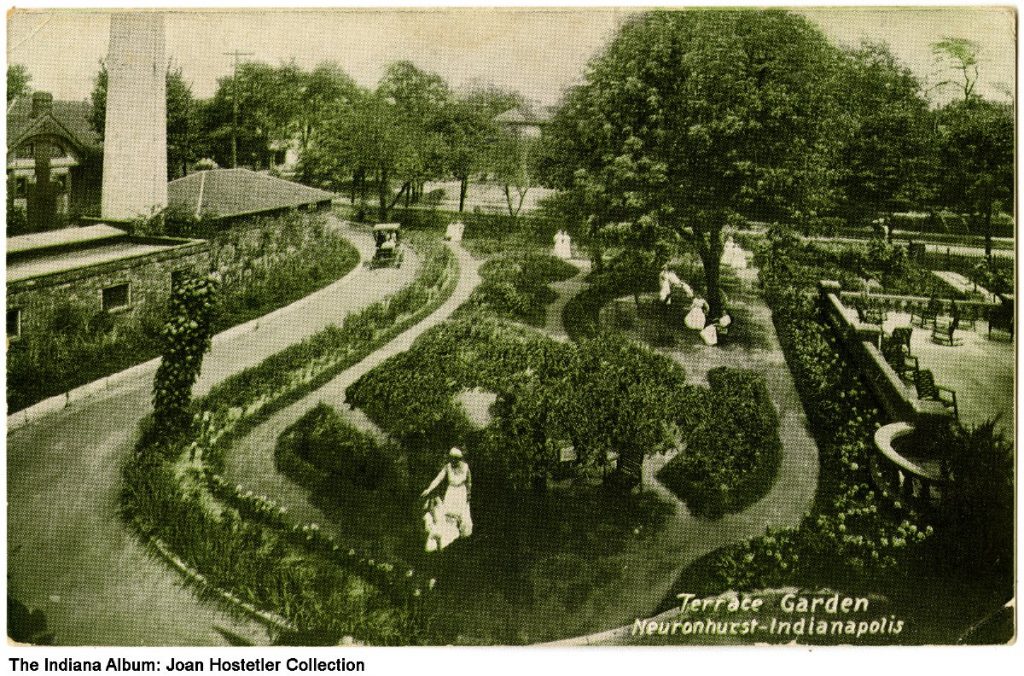 A large garden with several winding paths running through it. Several nurses walk and wheel patients around the garden. 