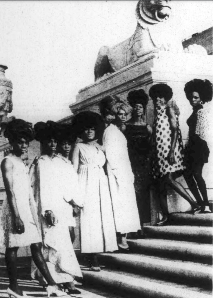 A line of eight people dressed in drag stand on the steps of a building. 