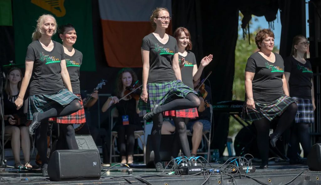 Two rows of dancers perform on stage. The dancers are wearing plaid skirts and t shirts that read Celtic Motion. 