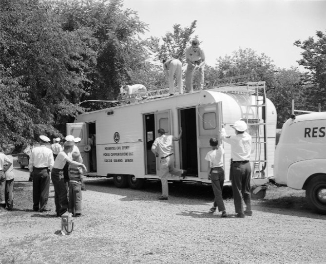 A group of people stand outside a van. The van reads "Indianapolis Civil Defense Mobile Communications Unit".