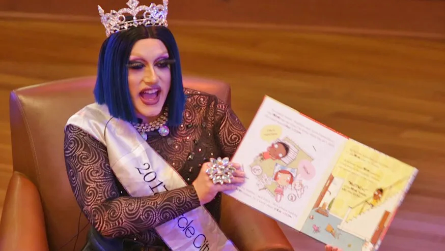 A person in drag dress sits in a chair on a stage and holds up a book. 