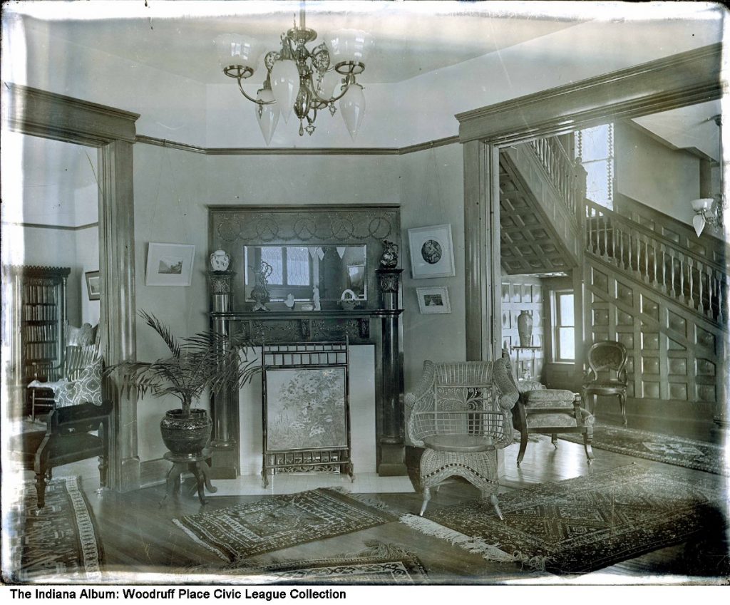 A view showing the fireplace in a corner of an ornately decorated parlor. There are entryways on either side of the fireplace. One leads to another room with chairs and bookshelves. The other leads to a hallway and staircase.