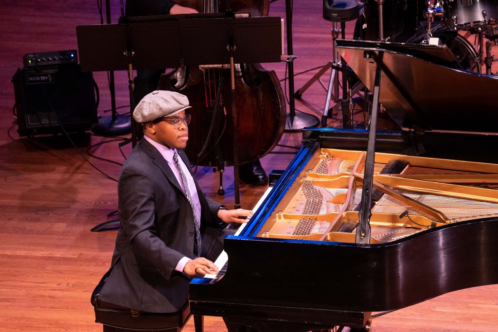 A young Black man sits at a piano on a stage. 