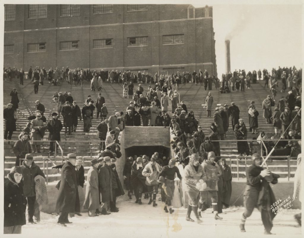 Several men in football uniforms emerge from an opening in the stands of a stadium. Spectators sit in the stands and stand on the ground outside the opening. 