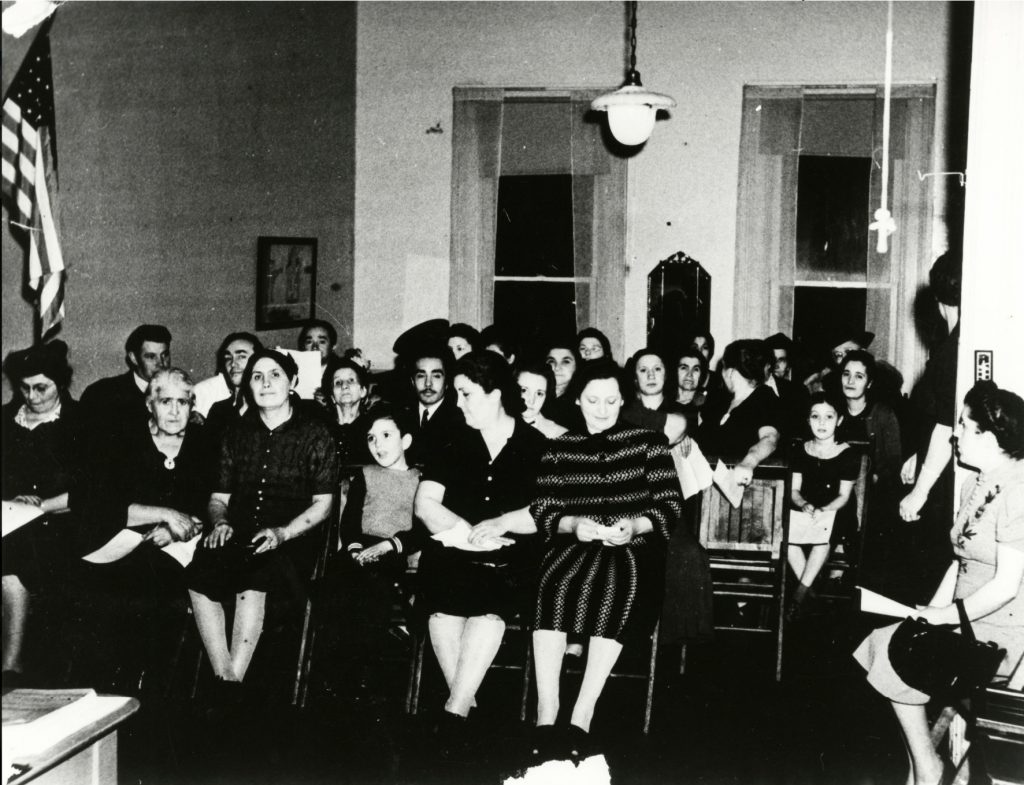 A group of people sit in rows of chairs facing the front of a classroom.