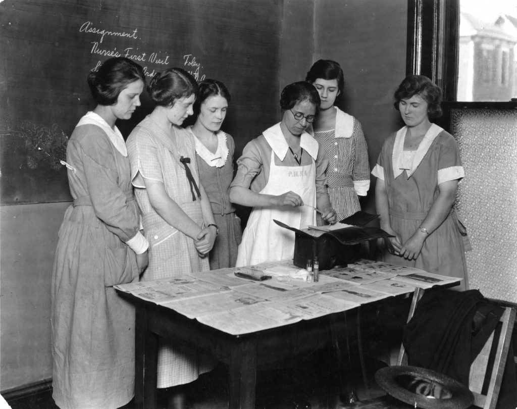 A nurse stands in front of a newspaper covered table and demonstrates a procedure. Five young women stand around the nurse to observe the procedure.