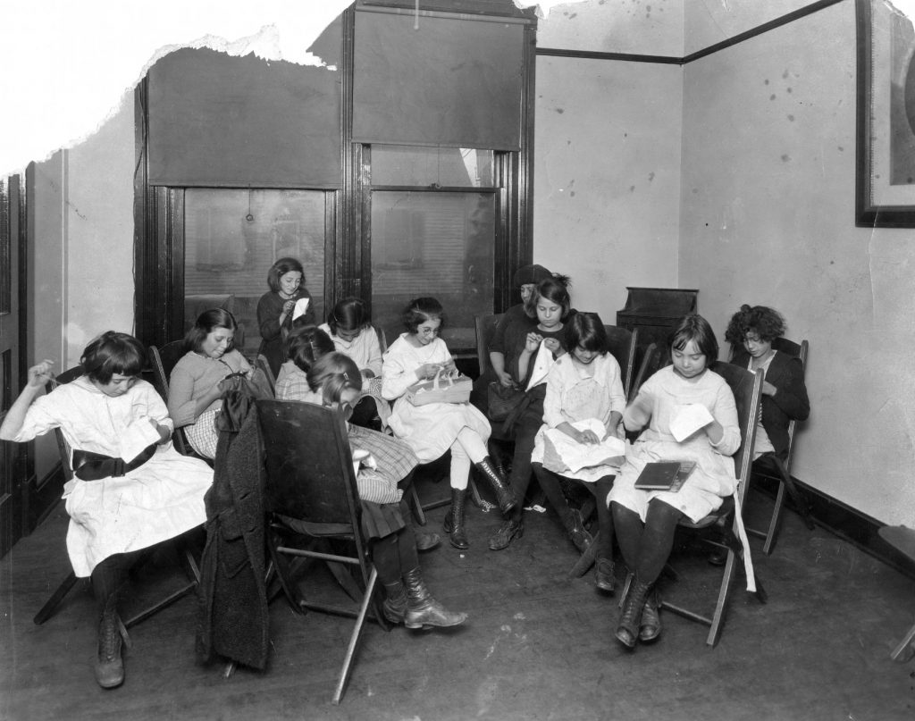 A group of 11 young girls sit together in a small room. Each girl is working on a sewing project. An older woman watches the girl's progress from the back of the room.