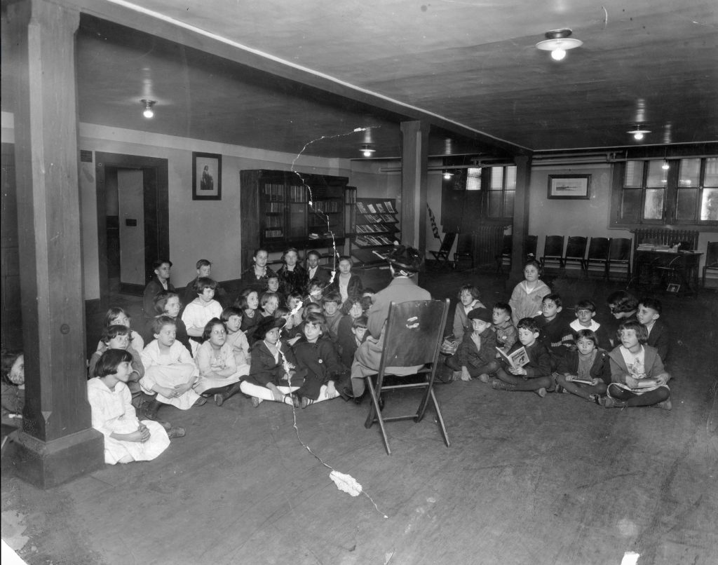 A group of children sit on the floor of a large open room. A woman sits in a chair in the middle of the children.