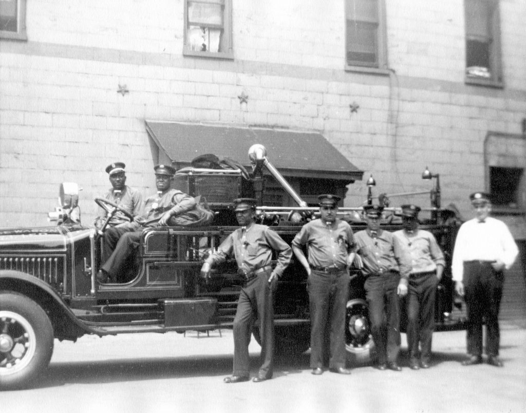 A fire truck is parked in front of a building. Five Black men stand in front of the truck and two Black men sit in the driver and passenger seat of the truck. 