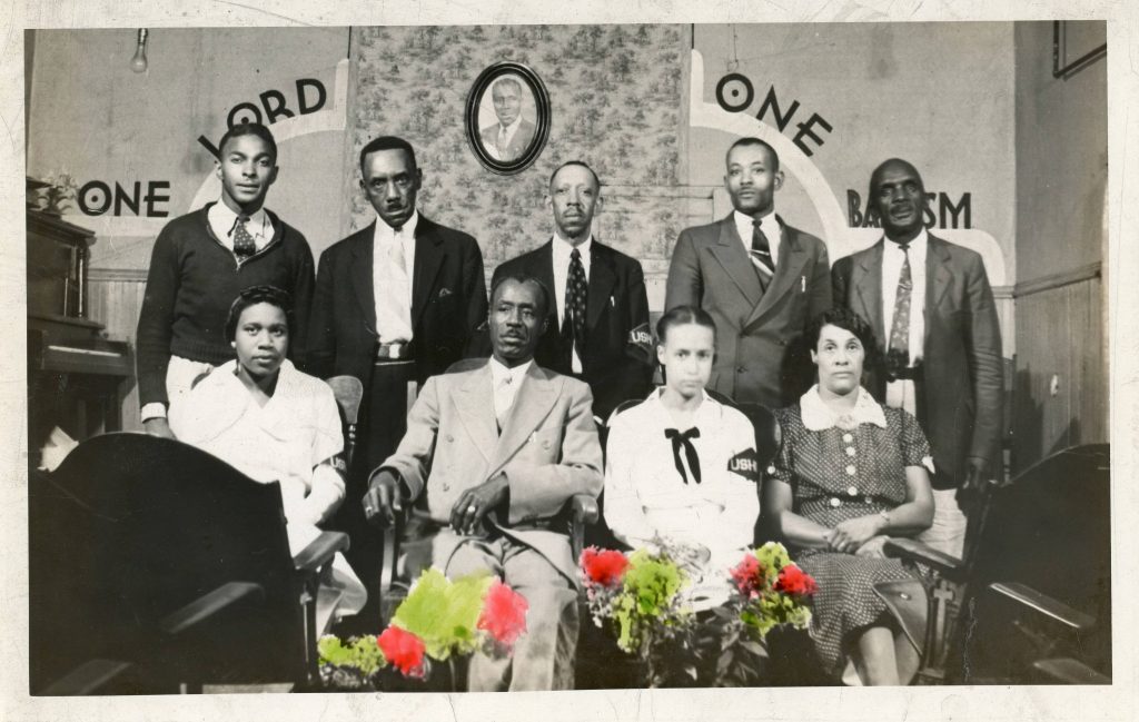 Nine people pose together for a photograph. Five men stand in back and one  man and three women are seated in front. Words painted on the wall in the background read "One Lord, One Baptism".