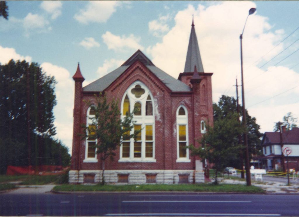 Exterior view of a red brick church building with ornate arched windows and a single spire.