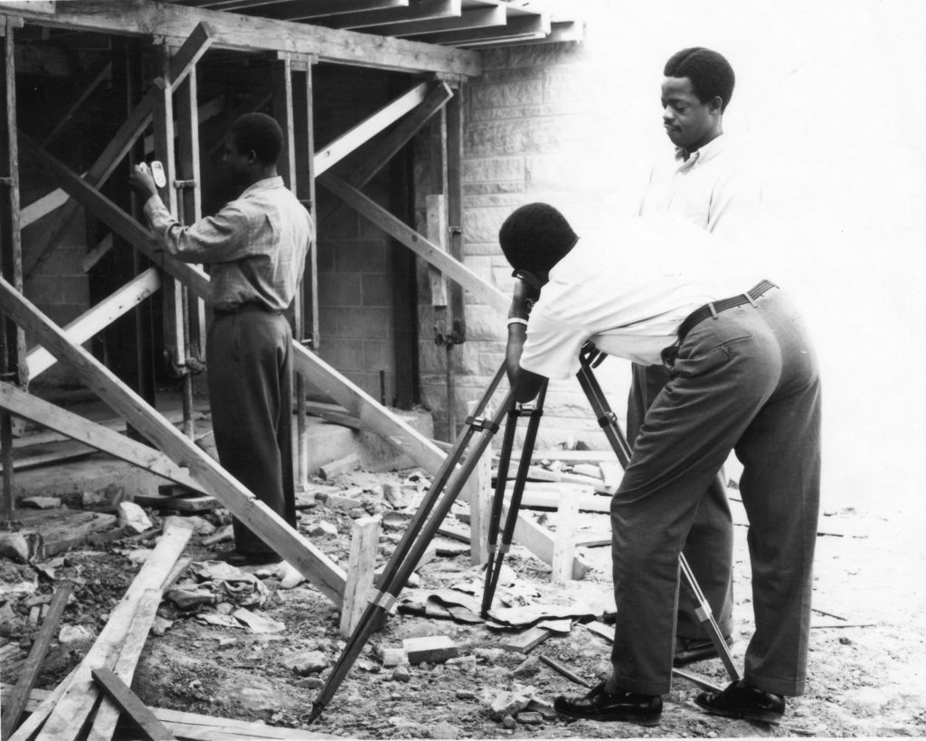 Three men stand at a construction site of a single story home. One is holding up a small tool, while another is using a tool attached to a tripod.