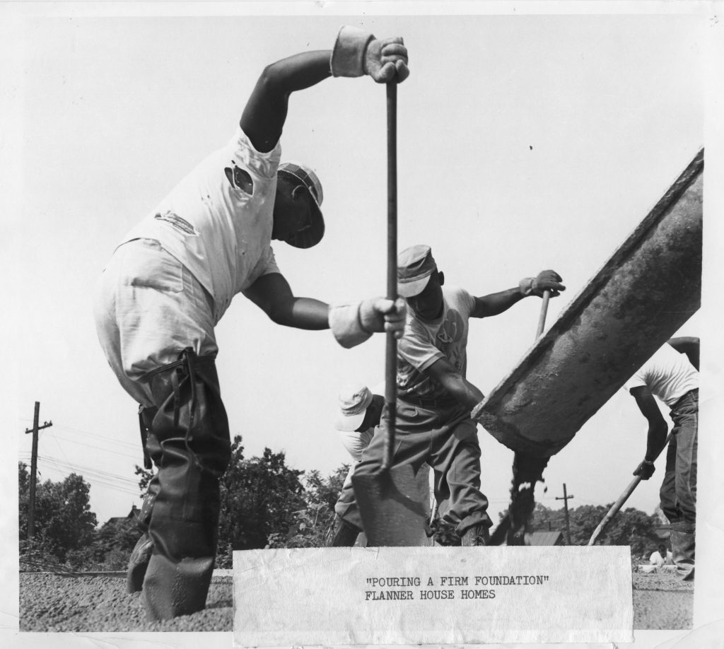 Four men in waders work to spread concrete for the foundation of a house. Concrete is actively flowing from a chute in the foreground.