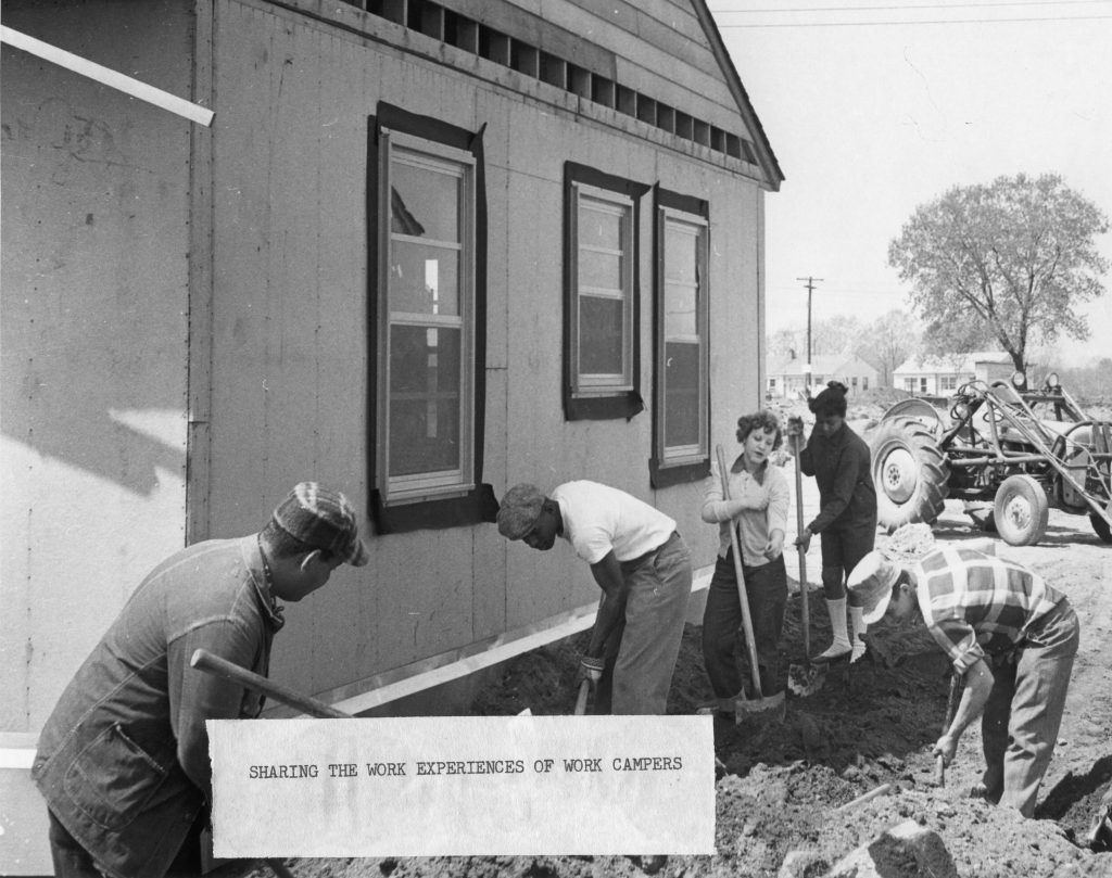 Five people all use shovels to level dirt around the foundation of a partially constructed single story home. Two of the people are young women.