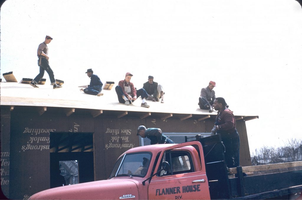 Several men are working on top of the roof of a partially constructed single story home. A truck is parked in front of the home and two men are standing in the bed of the truck.