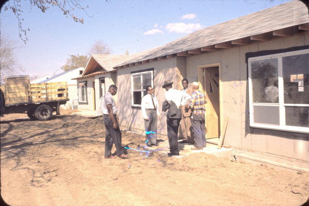 Five men stand outside the front step of a partially constructed single story home. The home next to it is also in mid-construction and the ground in front of the homes is still dirt.