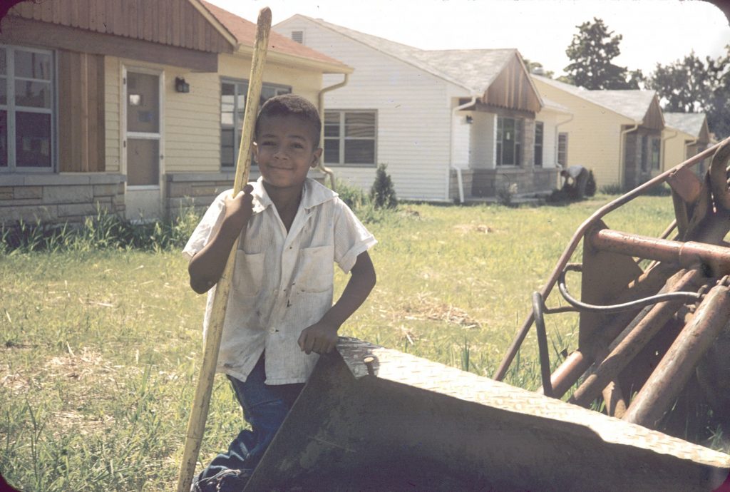 A young boy stands next to a large digger and holds a shovel. A row of newly built houses line the background.