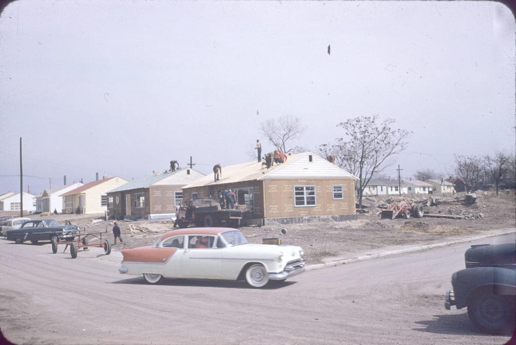 A row of five small single story houses. The two houses in the foreground are mid-construction and workers on standing on top of both roofs.