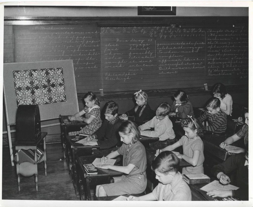 Students sit at individual desks in a classroom. All students are writing on a sheet of paper.