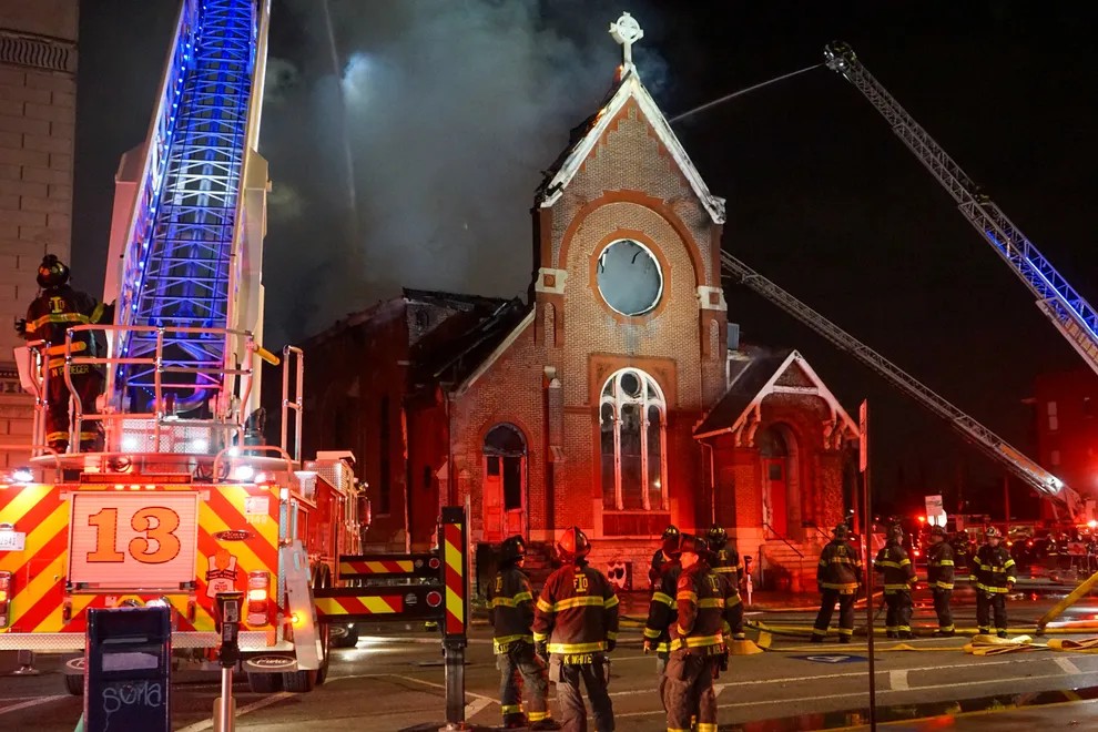 Firefighters stand on the street in front of a smoking church. One fire truck is in the foreground and the cranes of two other fire trucks can be seen. All cranes are spraying water on the building. It is night.