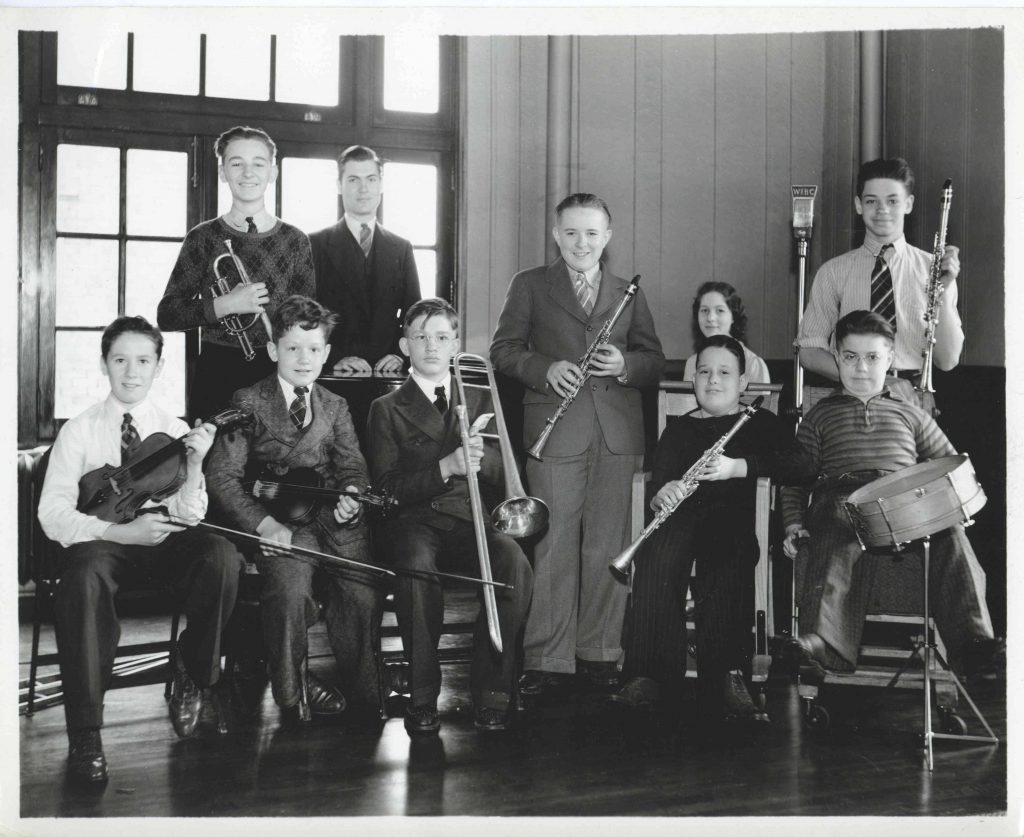 A group of students sit and stand next to each other. All are holding or playing an instrument. Two students are in wheel chairs.