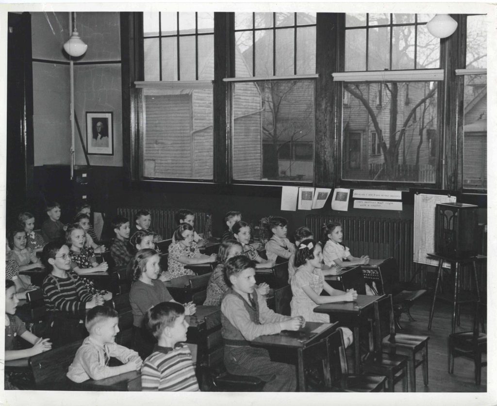 A group of young children sit at individual desks facing the front of a large classroom with several windows lining one wall.