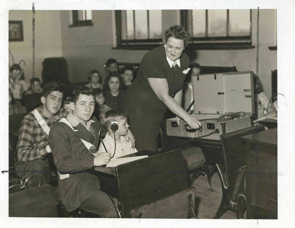 A teacher stands in the middle of a classroom and operates a turntable. Older students sit next to and help younger students. The younger students are wearing headsets and the older students are wearing a white sash with a badge on it.