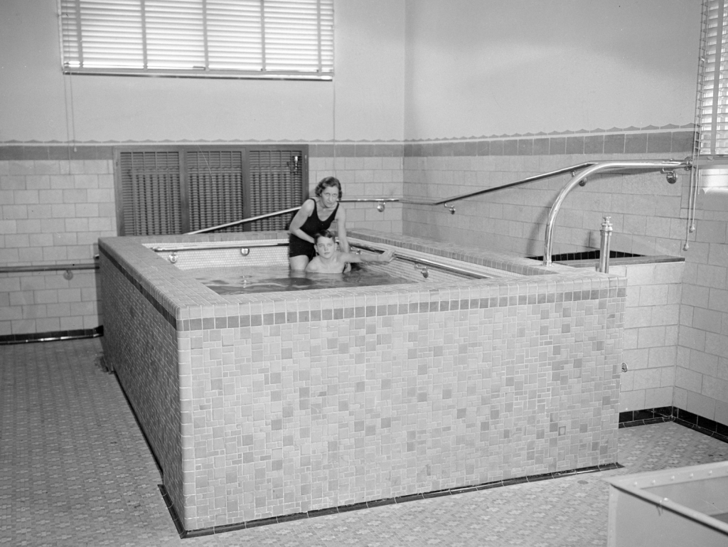 This interior photo of the James E. Roberts School shows a woman, possibly a therapist of teacher, helping a student in a hydrotherapy pool.