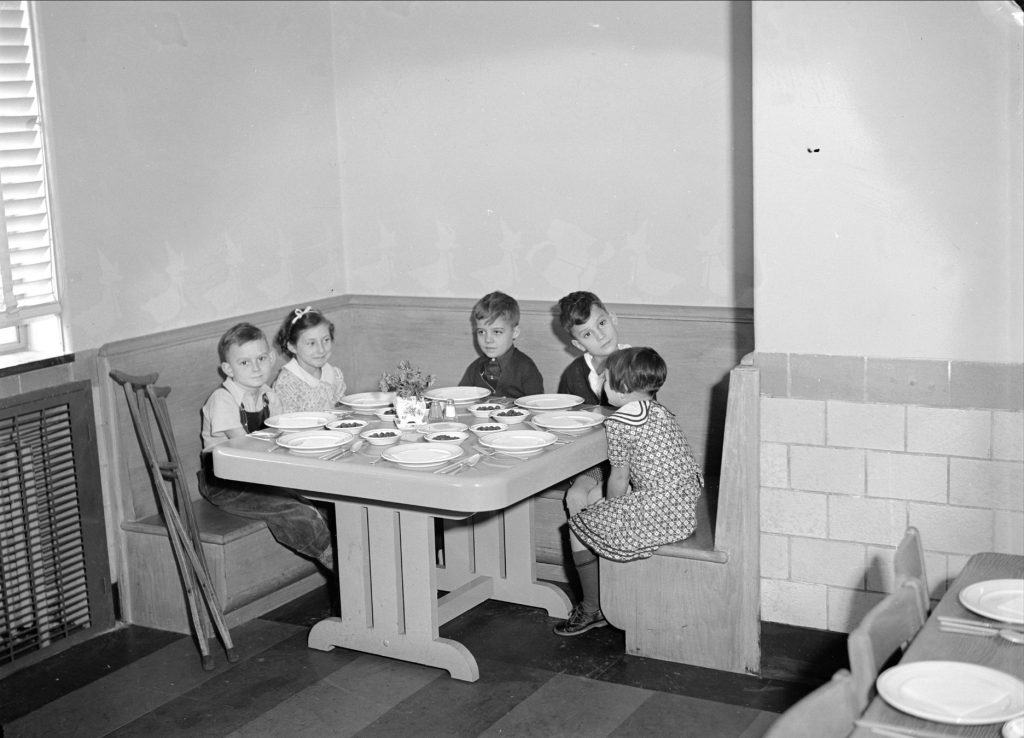 This interior photo of the James E. Roberts School shows five children at a table that is set for a meal. A set of crutches are propped up against a wall next to one student.