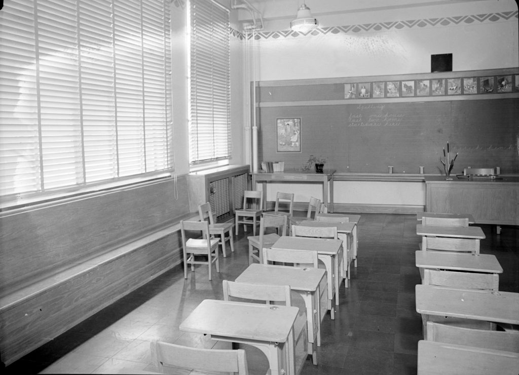 This interior photo of the James E. Roberts School shows a classroom with desks and blackboard. A row of windows fills one wall.