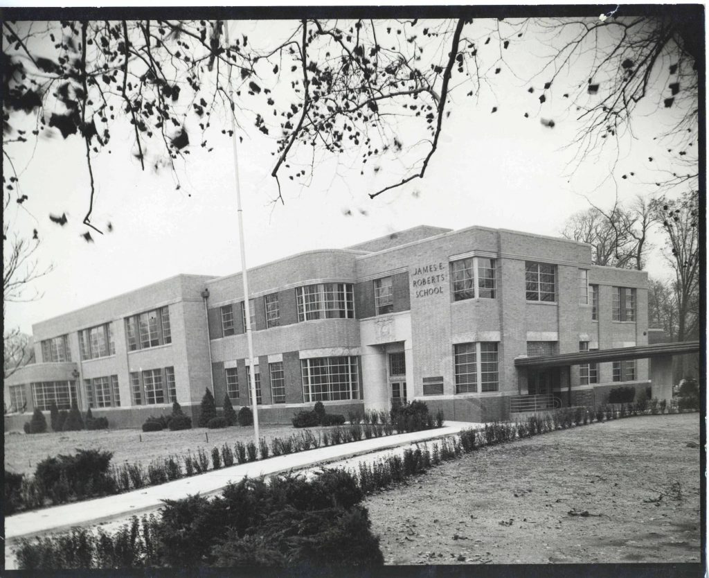 Far view of the front of a multi-story brick school building. The style is modern with squared windows and a curved wall protruding from the far side of the double entrance doors. Small shrubs line the walkway leading to the front entrance.