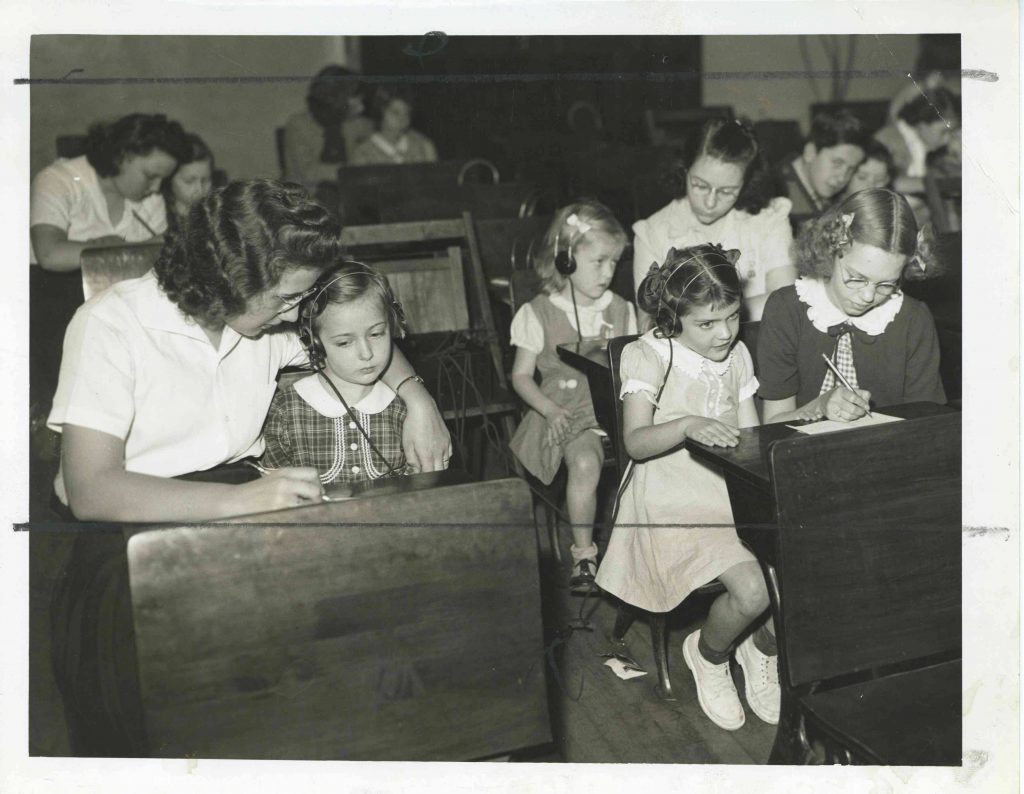 Teachers and older students each sit next to a younger student at desks. The younger students are all wearing headphones while the older students write on a sheet of paper.