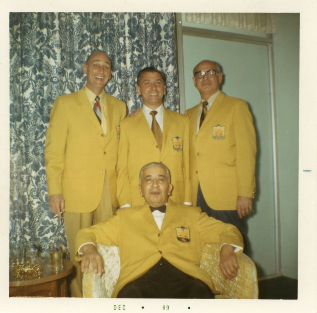 Four older men pose together in matching yellow jackets. Three of the men are standing and one man is sitting in a chair in front of them.