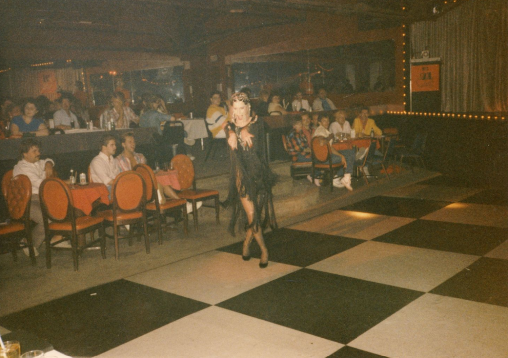 A drag performer dances in the middle of a dance floor while an audience of people watch from the side.