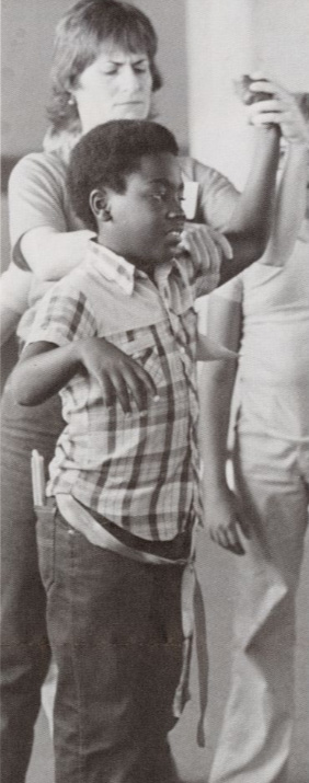 An adult positions the arm of a standing child in some kind of movement exercise.
