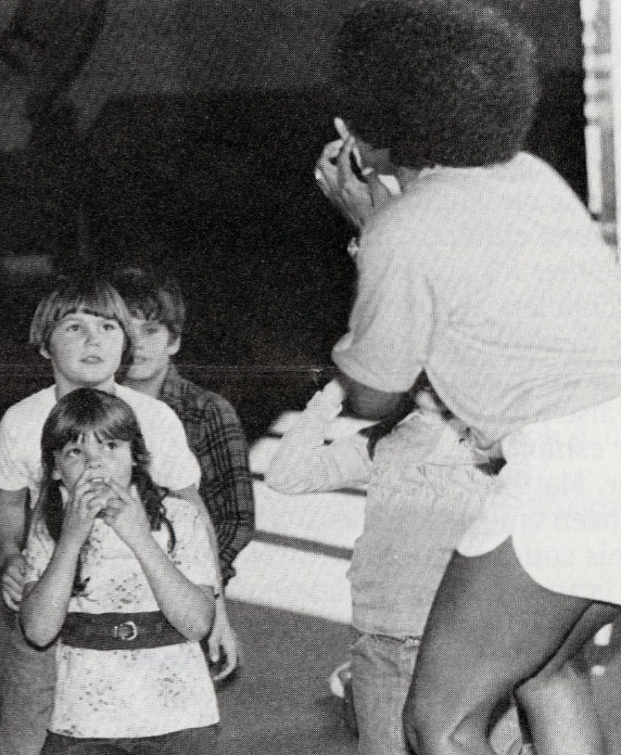 An adult stands in front of a group of children. She points to her face.