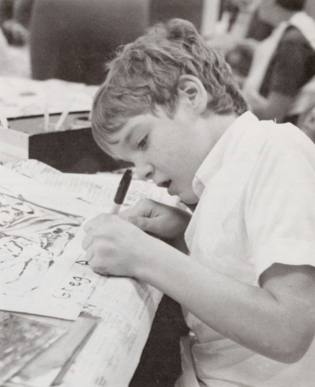 A child sits at a table and is writing his name on the bottom of a sheet of paper with drawings on it.