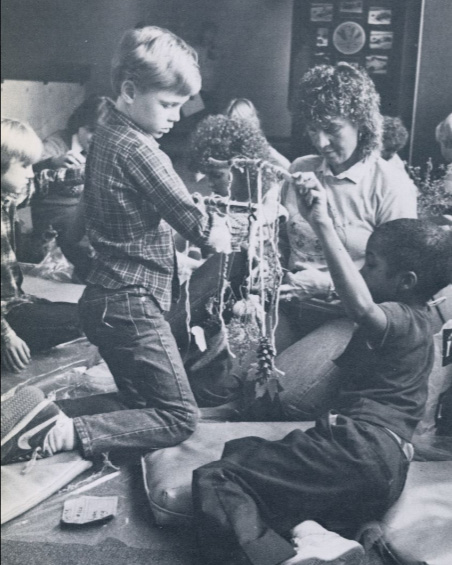 An adult sits in the middle of a group of children. Two children in the foreground hold up sticks with pine cones and other objects attached to them with strings.