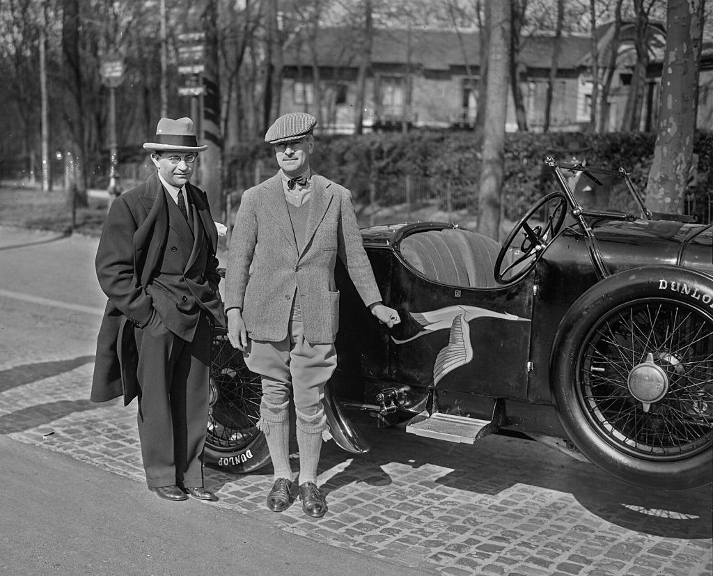 Two men stand next to an open top car.