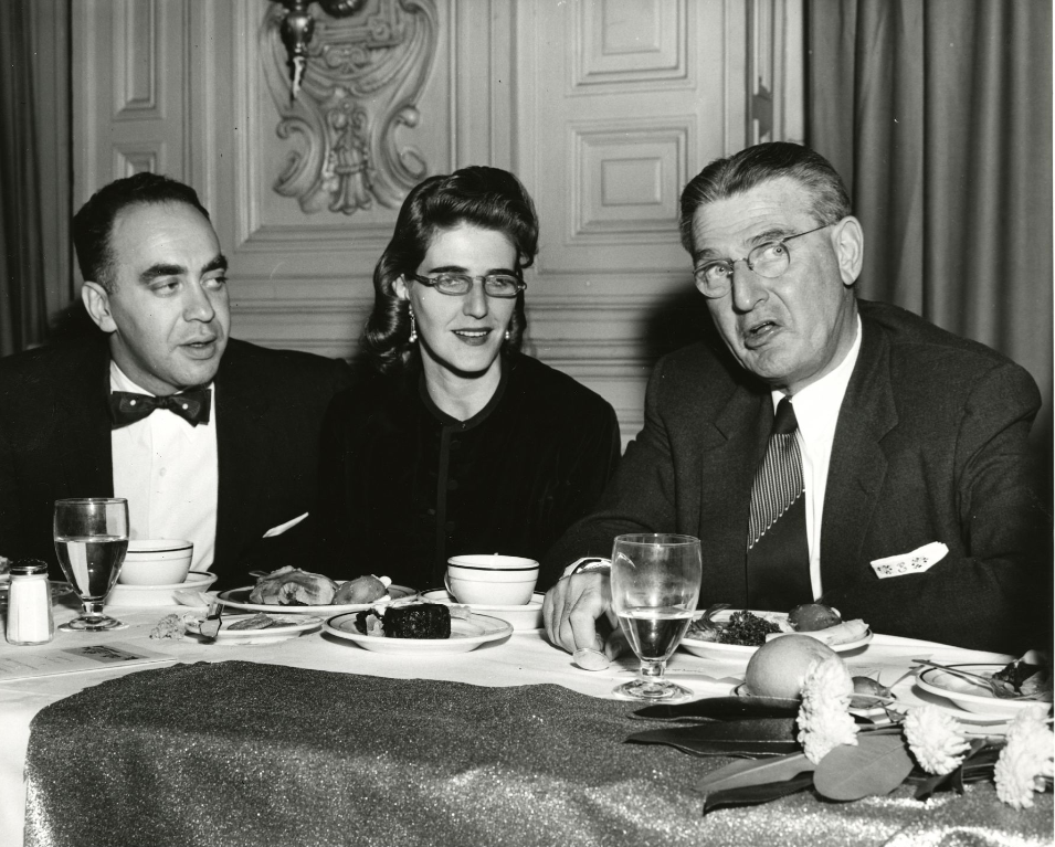 A middle-aged man and woman sit next to an older man at a table. Plates and bowls of food are in front of them.
